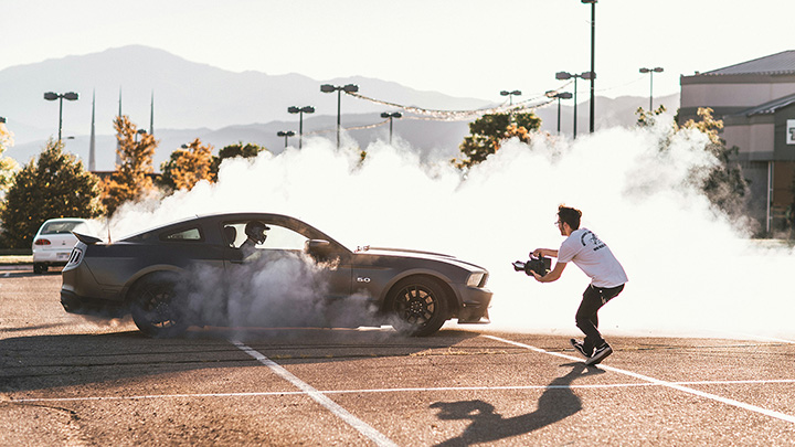 Person filming a car performing a tyre burnout in a car park, with smoke surrounding the vehicle.