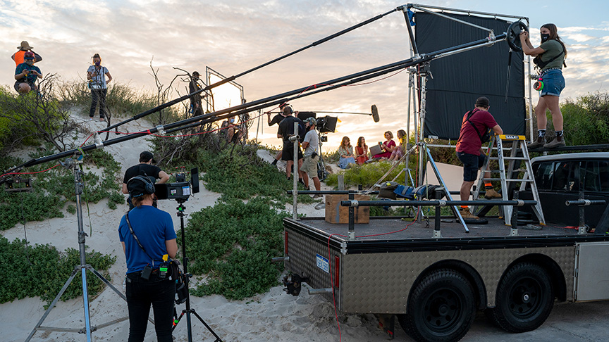 Film crew producing an outdoor scene on coastal dunes, using cameras, lighting, audio and rigging equipment.