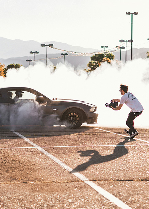 Person filming a car performing a tyre burnout in a car park, with smoke surrounding the vehicle.