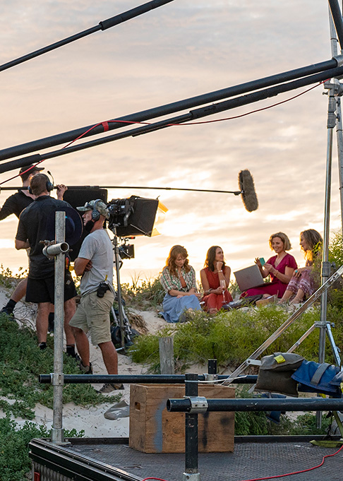 Film crew recording a group discussion outdoors on coastal dunes at sunset, with cameras, lighting and audio equipment visible.