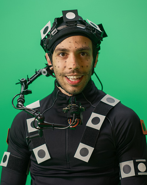 Person wearing a motion capture suit and head-mounted camera rig in front of a green screen.